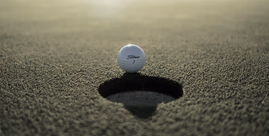 A golf ball with the brand Titleist and the number 3 rests on the edge of a golf hole on closely-cut grass.